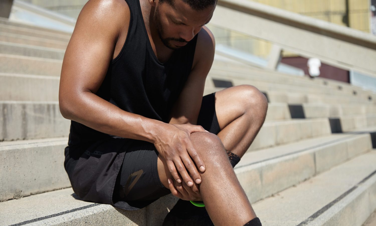 Handsome muscular male jogger wearing black training outfit touching his knee in pain with clasped hands, having sprain or rupture in his muscles after exercising outdoors. Sports injury concept body dehydration