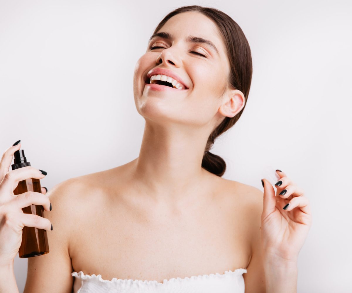 Satisfied brunette girl after shower sprays mist for body on skin. Woman smiling on isolated background. skincare